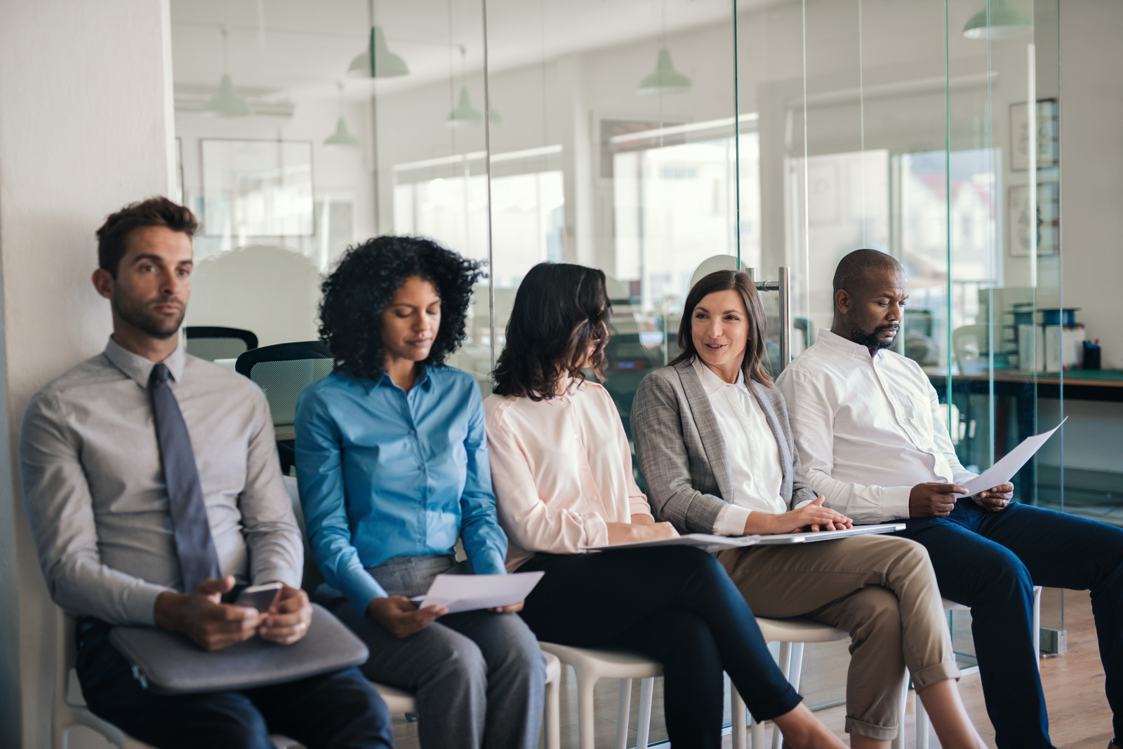 A group of people – three women and two men – wait outside an office for their interview holding CV printouts and laptops