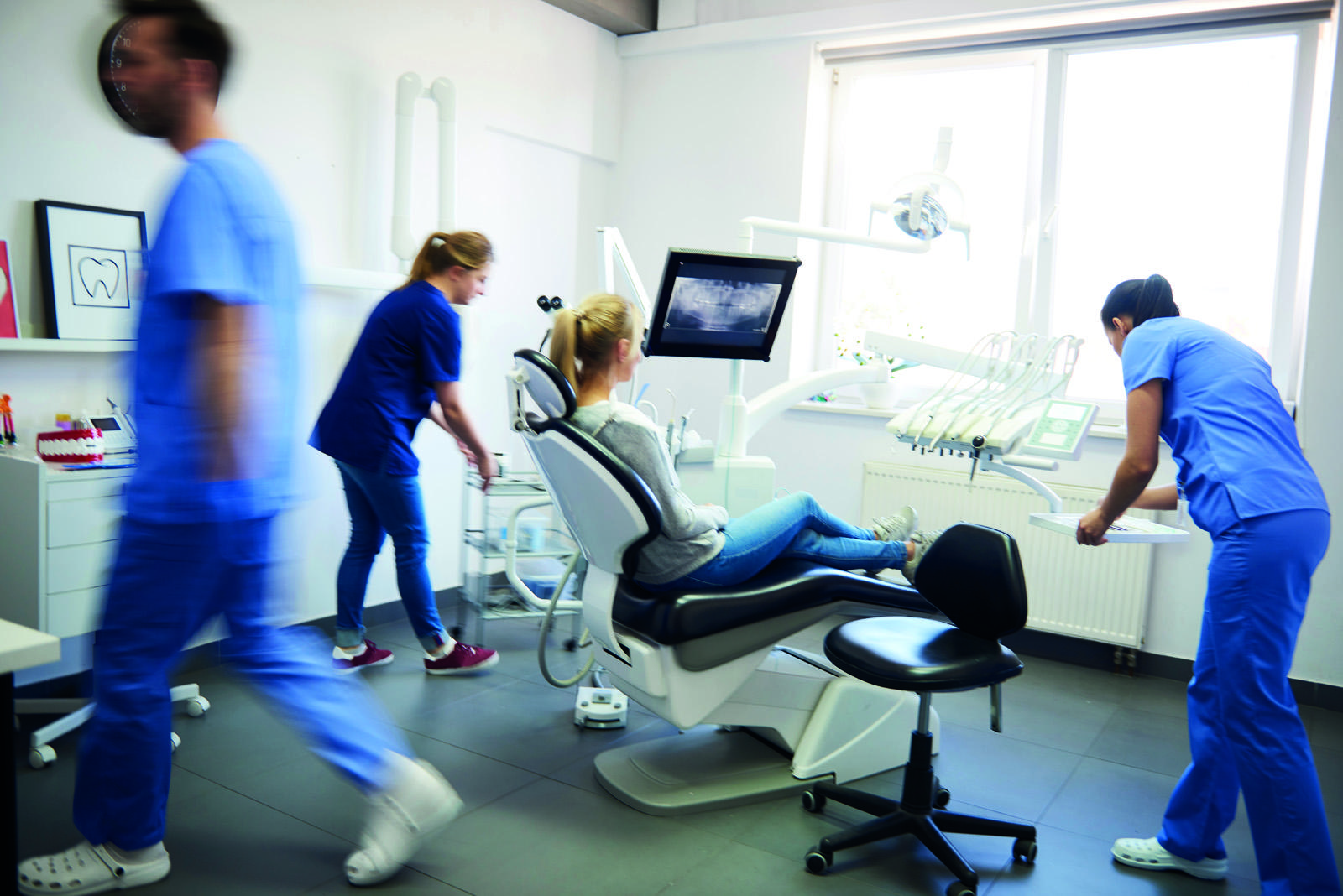 A busy dental team in surgery with a blonde patient in the chair facing away from the camera towards a bright window