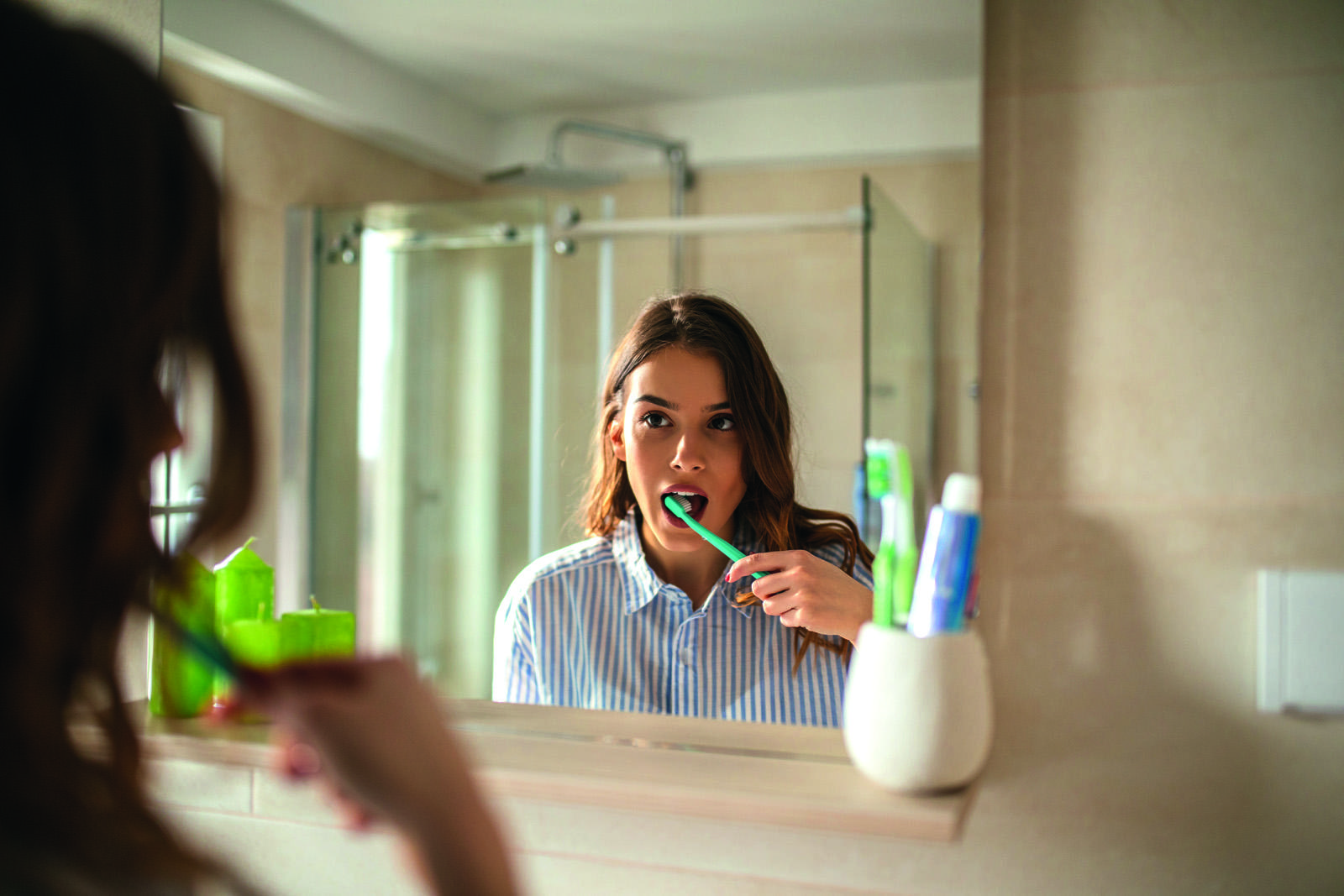 Young brunette woman brushing her teeth in a mirror wearing striped pyjamas in a sunny bathroom