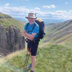 John Gibson on his walk leaning on his walking poles with a view down a beautiful green valley stretching away in the distance