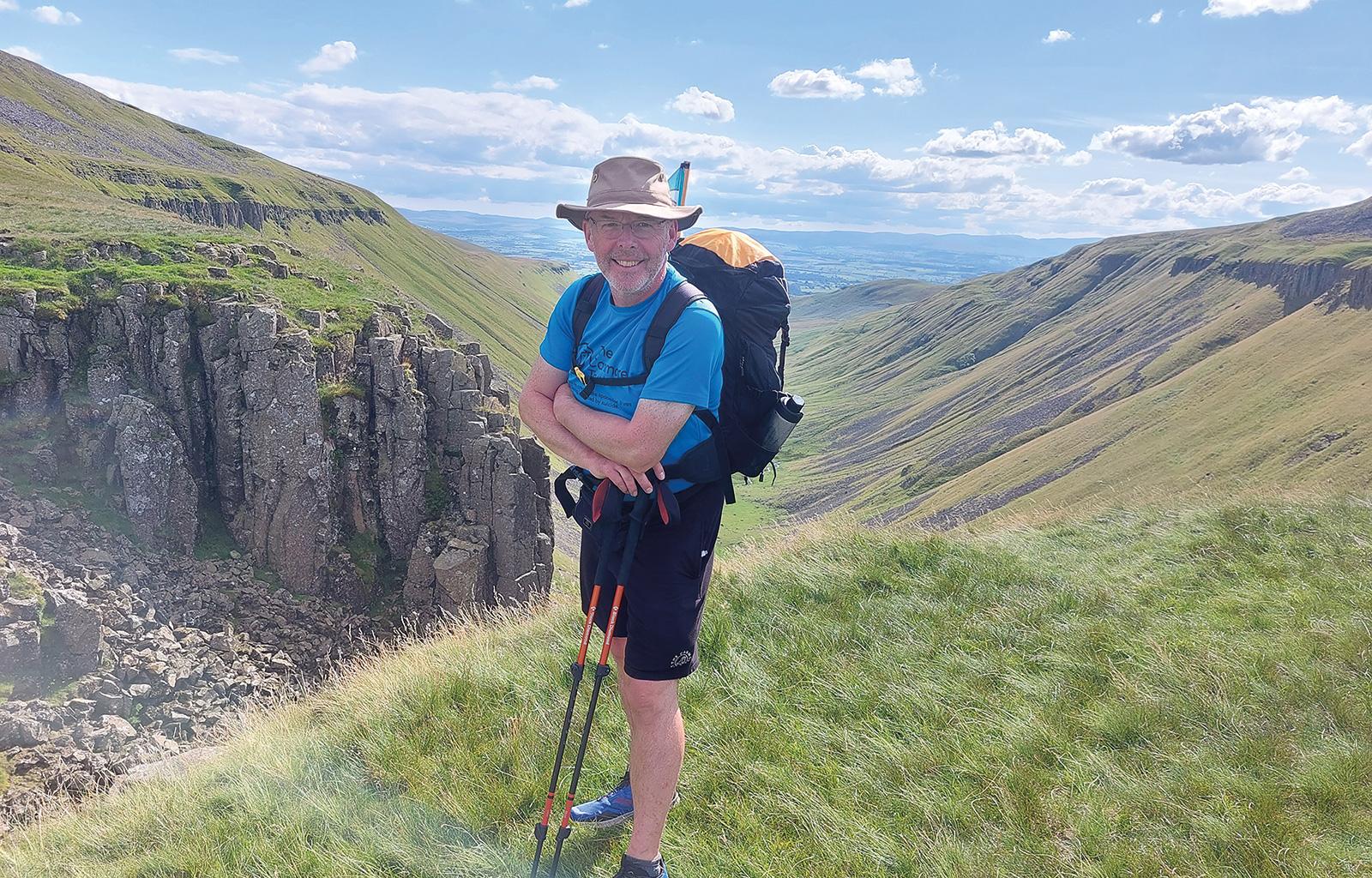 John Gibson on his walk leaning on his walking poles with a view down a beautiful green valley stretching away in the distance