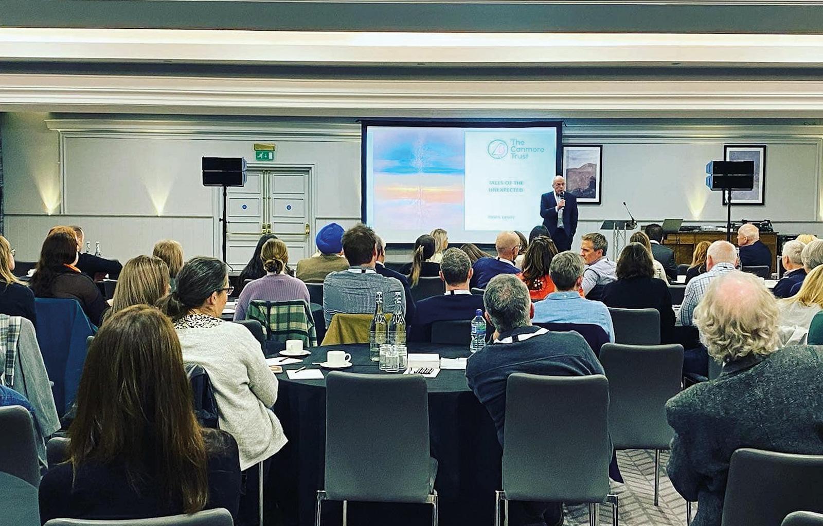 Professor Gibson Delivers a talk at the symposium, photographed from the back of a packed room on stage in front of a presentation screen